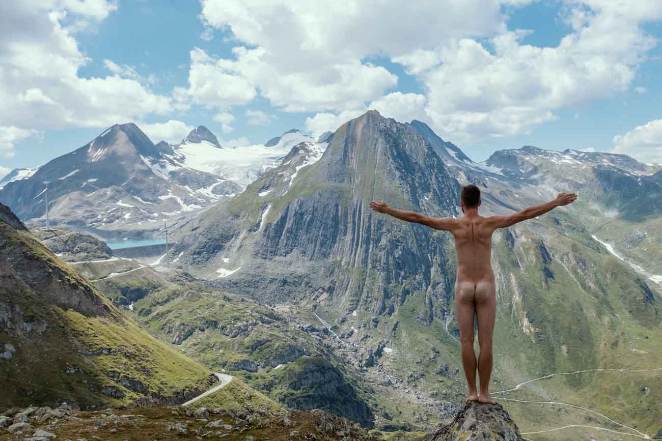 Patrick Arendt steht komplett nackt mit dem Rücken zur Kamera und guckt auf das Bergpanorama