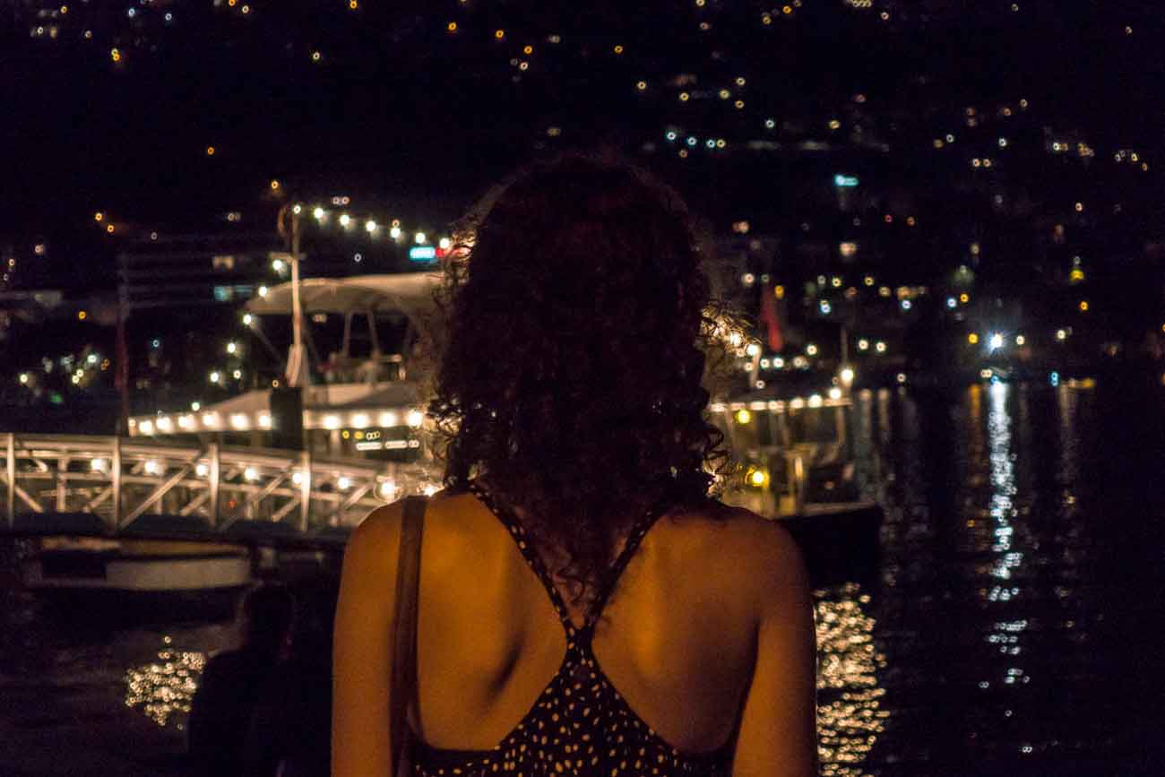 Eine Frau guckt auf den Lago Maggiore in der Nacht und steht mit dem Rücken zur Kamera. Auf dem Wasser liegt ein Boot und ist beleuchtet